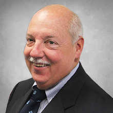 board member headshot wearing black suite with blue and white shirt with a navy blue tie