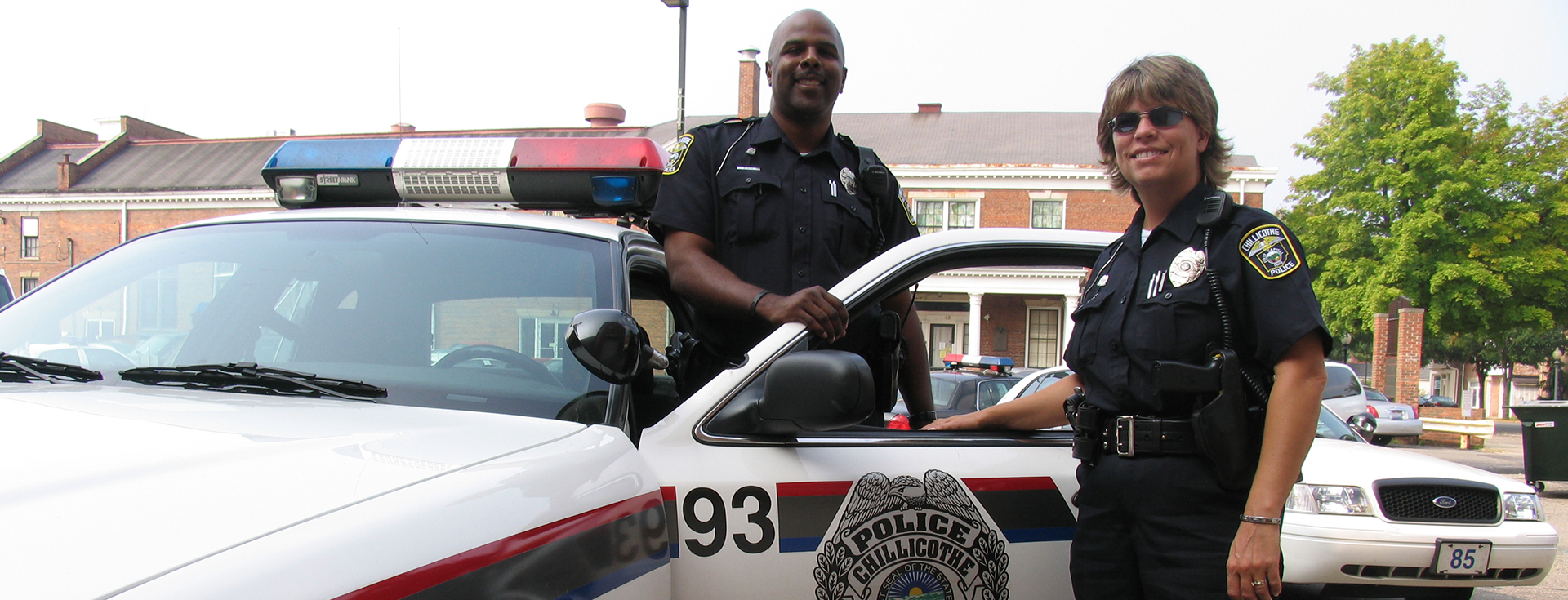 one male and one female police officer standing by a white with red and black striped police car with the driver side door open