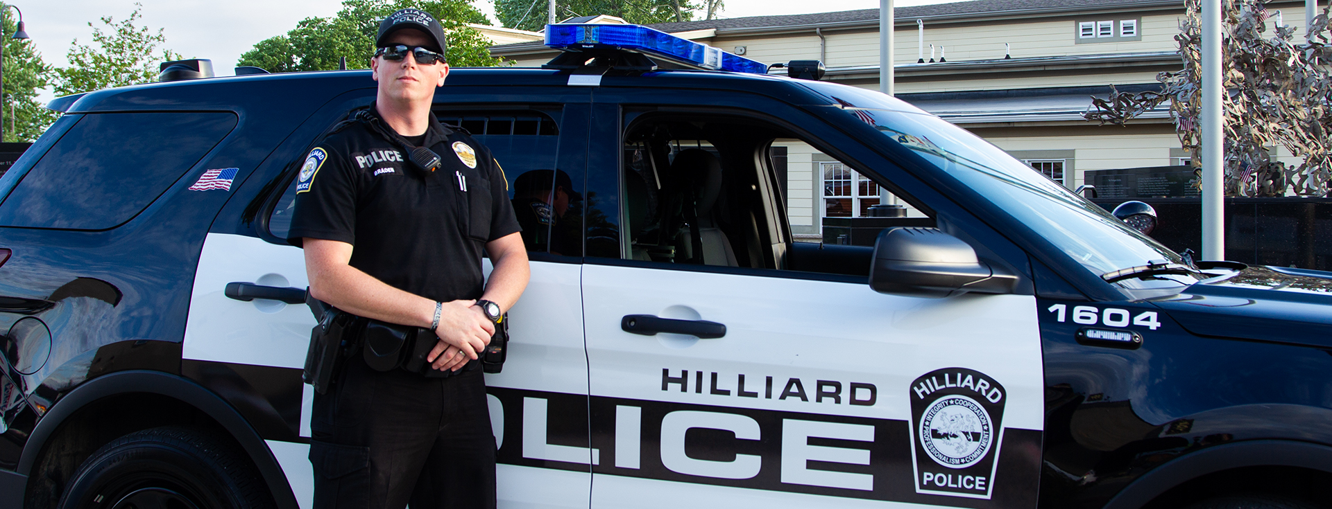 police officer wearing a hat and sunglasses standing with his arms crossed next to ta black and white police SUV vehicle